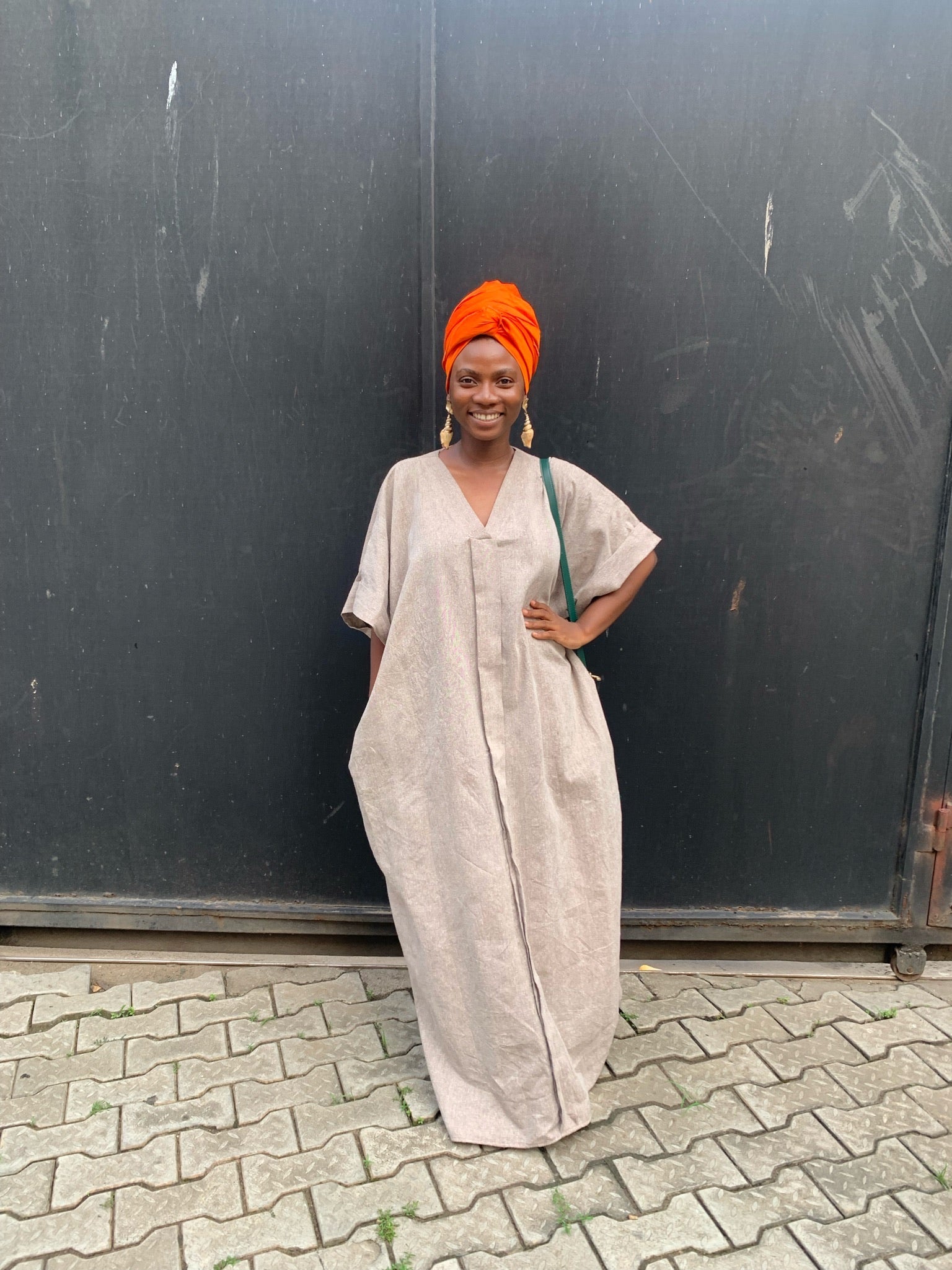 Woman in loose beige kaftan dress with orange headwrap, standing outdoors on paved ground