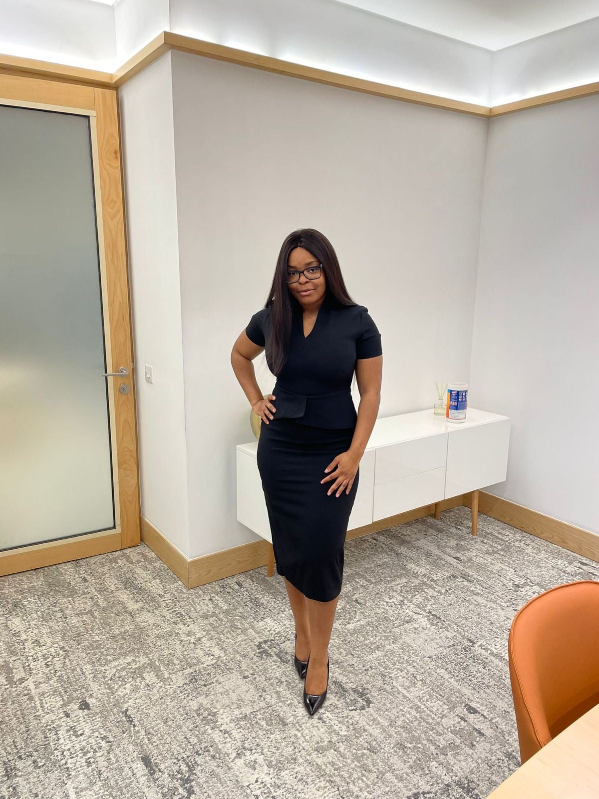 Woman in fitted black dress and heels posing confidently in a modern office setting