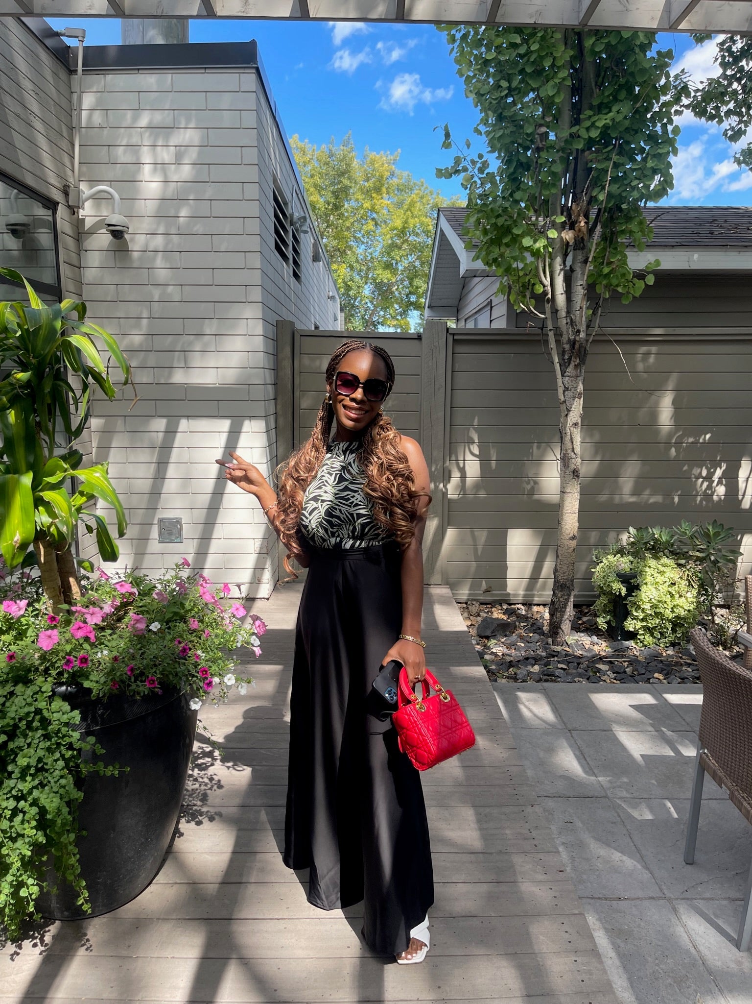 Woman wearing high-waisted wide-leg black pants with patterned top, outdoors in a garden setting, holding a red handbag.