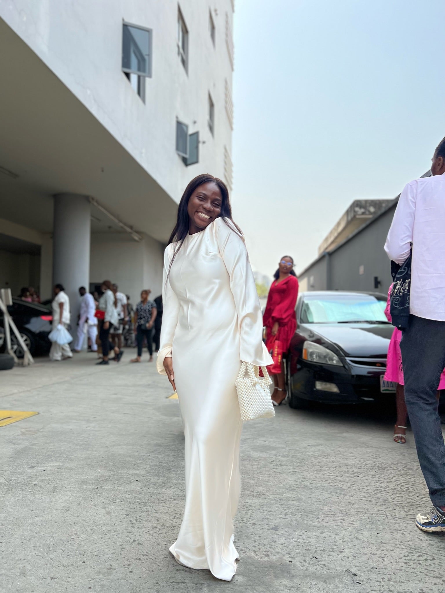 Woman in elegant white satin dress holding pearl handbag outdoors, smiling in Lagos street fashion